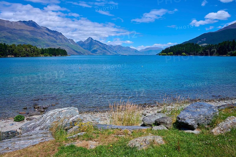 Tranquil seascape with grassy rocks and mountains in the background, Queenstown, Queenstown-Lakes District, Otago, South Island, New Zealand