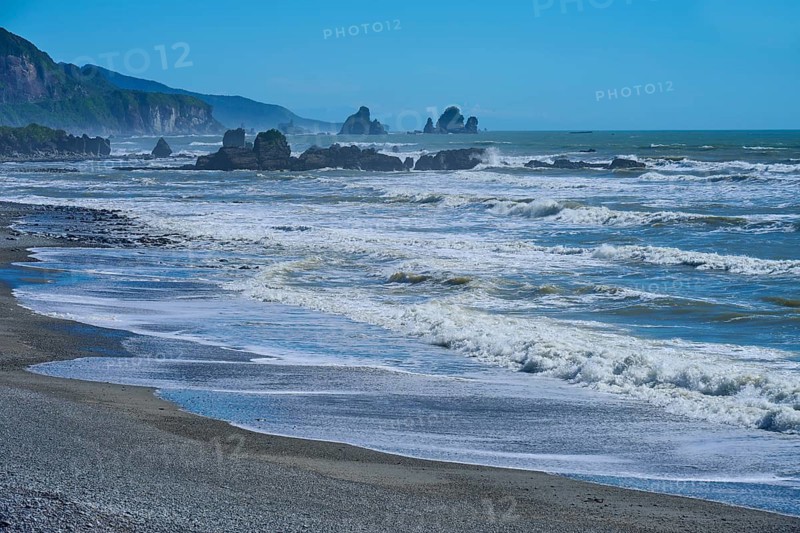 Dynamic waves roll onto a rocky beach under a bright blue sky, West Coast, South Island, New Zealand