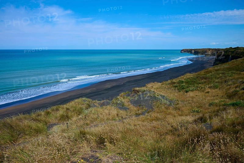 View of black sand beach with gentle waves, grassy coastline under clear sky, Mana Bay, Riverview Lookout, Patea, Taranaki, North Island, New Zealand