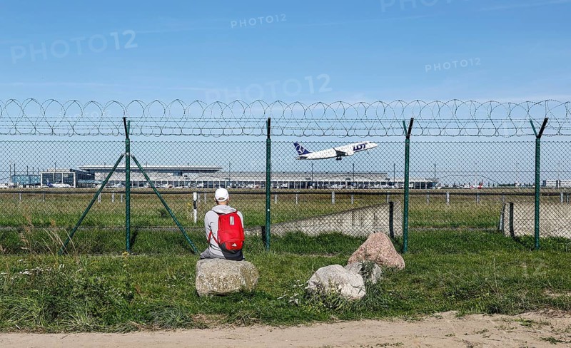 A man sits on a stone and watches an aircraft of the Polish airline LOT take off through a barbed wire fence at BER Airport, Berlin Schönefeld, 28/09/2025, Schönefeld, Brandenburg, Germany