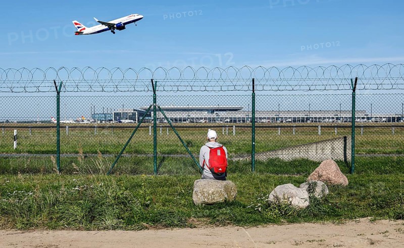 A man sits on a stone and watches the take-off of a British Airways plane through a barbed wire fence at BER Airport, Berlin Schönefeld, 28/09/2025, Schönefeld, Brandenburg, Germany