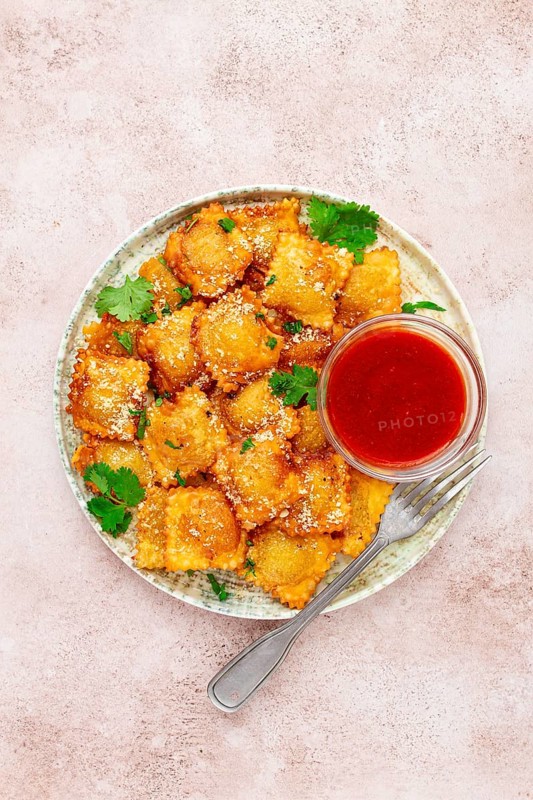 Fried ravioli with parmesan cheese, served with red sauce and garnished with herbs, on a light background, no people
