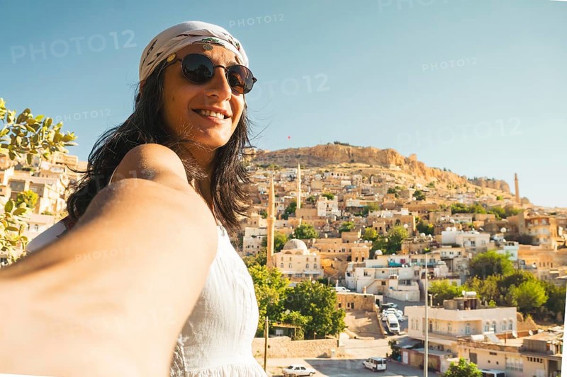 Smiling woman in sunglasses taking selfie with panoramic skyline of historic Mardin Turkey and mosque minarets on Mesopotamian hillside