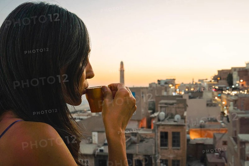 Tourist woman enjoying traditional Mirra coffee on rooftop with Mardin skyline and Mesopotamian plains at sunset, traditional Turkish Arab culture