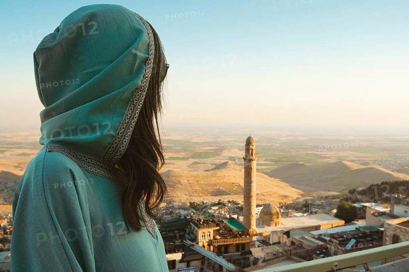 Smiling woman traveler in abaya standing above Mardin Turkey with panoramic view of Mesopotamian plains and historic mosque minaret at sunset