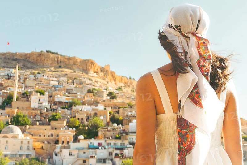 Traveler woman in white dress looking over panoramic view of historic city of Mardin Turkey with mosque minarets and traditional houses and Mardin castle on hilltop
