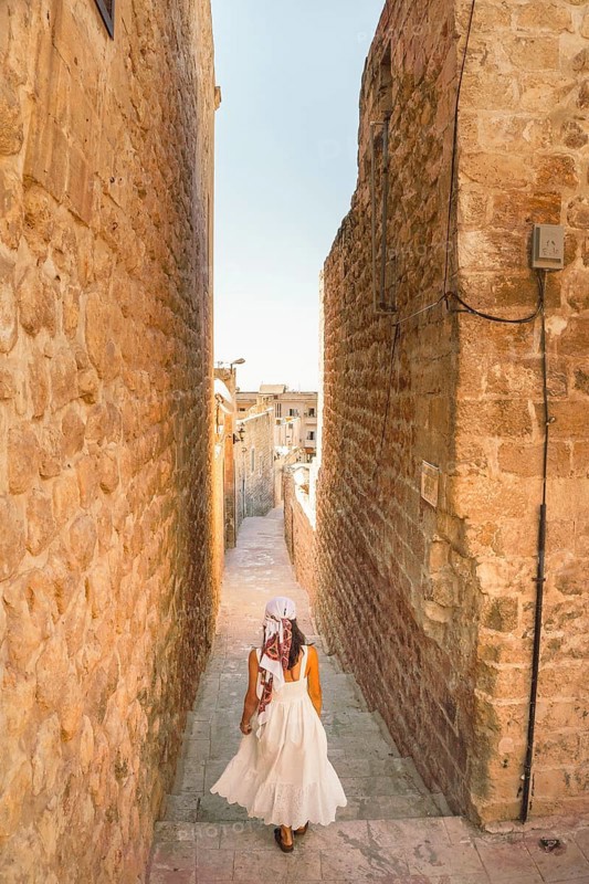 Traveler woman in white dress walking through narrow stone alley in historic city of Mardin Turkey with traditional architecture