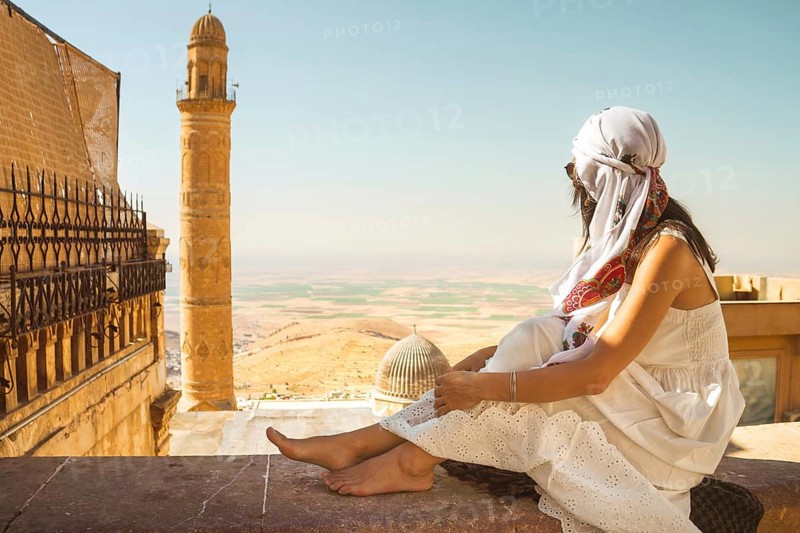Traveler woman in white dress sitting near Great Mosque of Mardin Ulu Camii with panoramic view of Mesopotamian plains and historic minaret