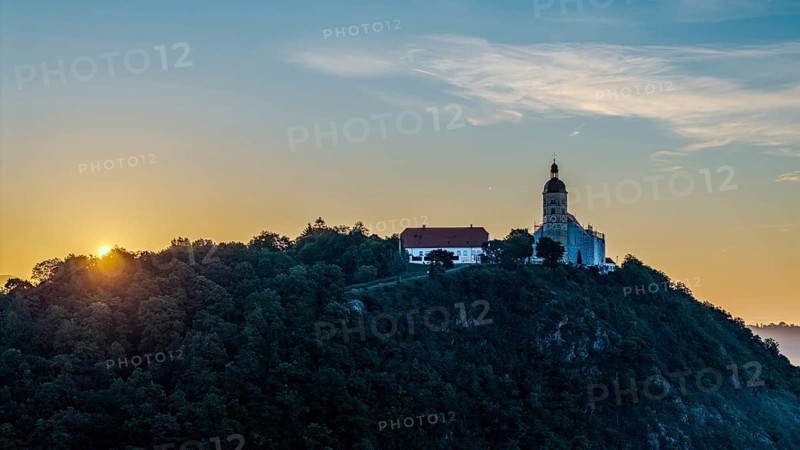 The sunrise illuminates the pilgrimage church of the Assumption of the Virgin Mary on the Bogenberg. The sky is tinged with soft colours. Trees surround the site and create a peaceful atmosphere. Bogen, Lower Bavaria