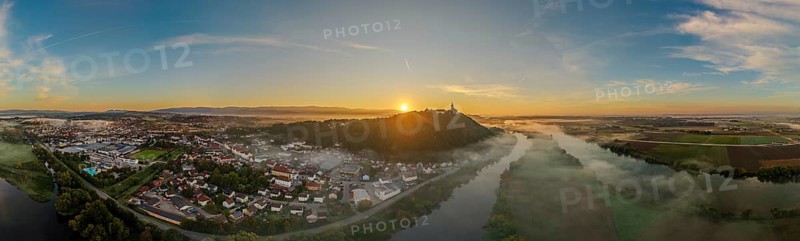 The sun rises over a city surrounded by fog. The picture shows the river landscape of the Danube and the Bogenberg with the historic pilgrimage church of the Assumption of the Virgin Mary in the background. Bogen, Lower Bavaria