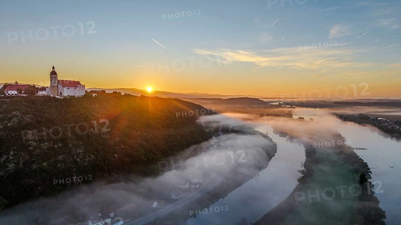 The sun rises over the Danube while mist drifts over the water. A tranquil landscape with trees and the historic pilgrimage church of the Assumption of the Virgin Mary on Bogenberg hill stretches out in the foreground. Bogen, Lower Bavaria