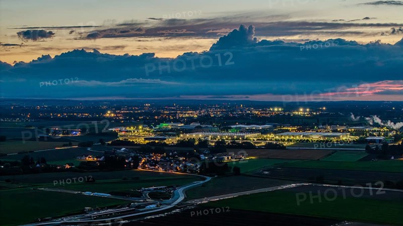 The scene shows an expansive landscape at dusk. Illuminated buildings and streets are visible in the distance, surrounded by clouds and rolling hills. Straubing, Lower Bavaria