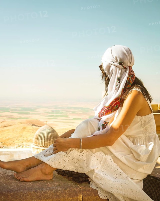 Traveler woman or tourist wear white dress sitting near Great Mosque of Mardin overlooking vast Mesopotamian plains with historic architecture in foreground