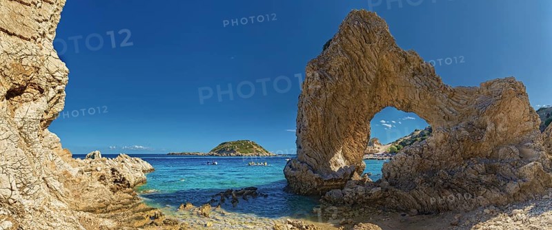 Stone arch on Grande Blue Beach in front of a small island and a rocky coastline under a clear blue sky, Grande Blue Beach, Stegna, Archangelos, Rhodes Island, Greece