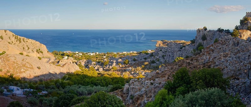 Coastal landscape of Stgna overlooking the sea, framed by rocky hills and lush vegetation, under a blue sky, Grande Blue Beach, Stegna, Archangelos, Rhodes Island, Greece