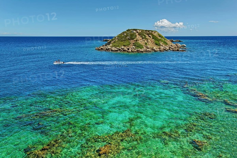 Small island Vrachonisida in front of the Grande Blue Beach in the turquoise blue sea with passing boat and blue sky with a cloud, Grande Blue Beach, Stegna, Rhodes Island, Greece
