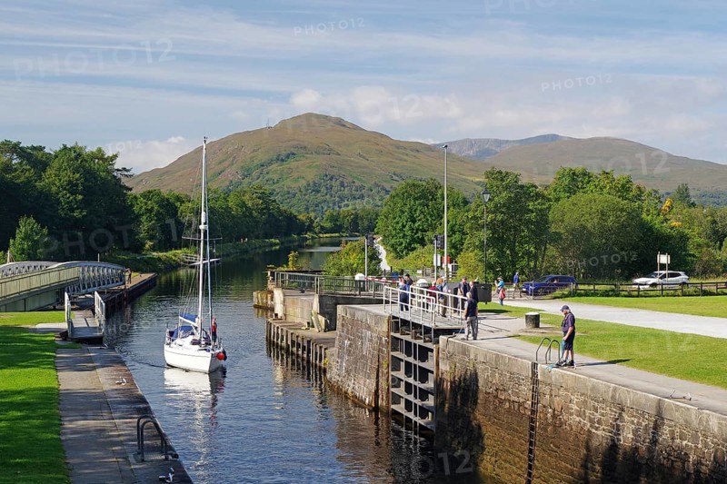 A sailing boat in the canal surrounded by green trees and mountains on a clear day, Caledonian Canal, Fort William, Scotland, United Kingdom