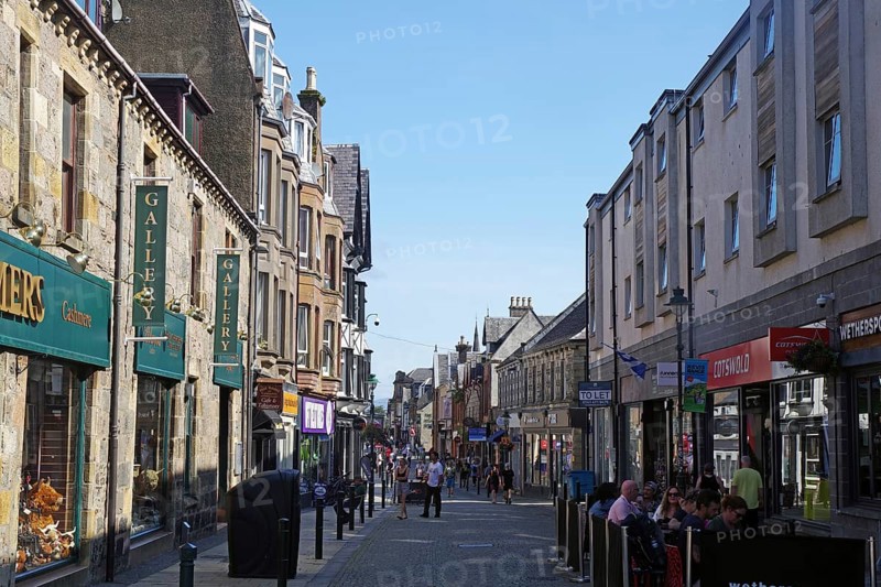 Busy shopping street with people and traditional buildings under a blue sky, Fort William, Scotland, United Kingdom