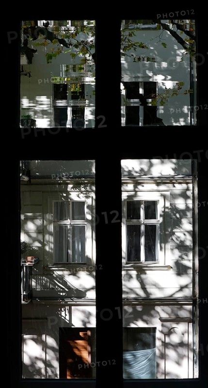 View through a window to an opposite facade of a multi-storey residential building with classical architecture and balconies with plants, shadows of leaves and branches on the house wall, Berlin, 24.09.2025, Berlin, Berlin, Germany