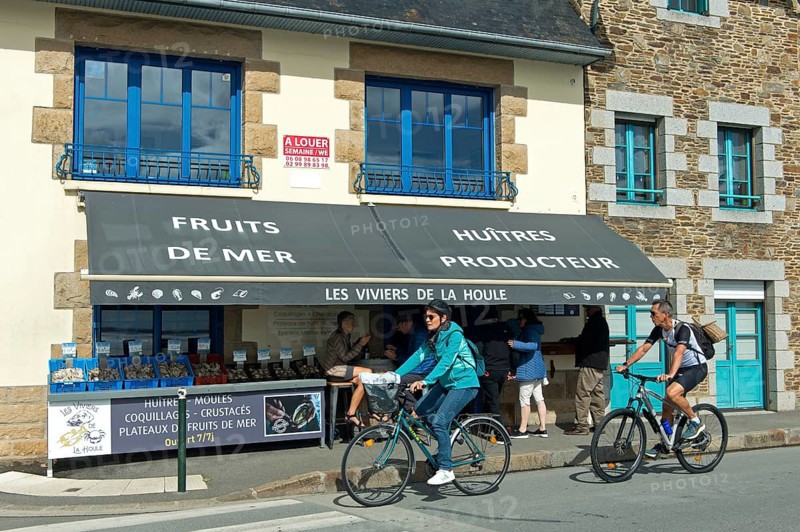 Cyclists on the Quai John Kennedy pass a shop selling seafood such as oysters and mussels, Cancale, Brittany, France