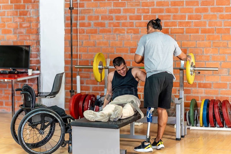 Man with prosthetic leg spotting another man with leg injuries or absence, who struggles with a barbell on a bench press, demonstrating dedication to overcoming challenges in a fitness setting