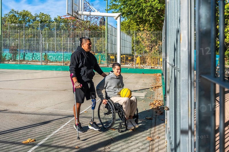 Friends celebrating sports inclusion on a sunny outdoor basketball court, one man with crutches and a prosthetic leg stands while pushing his smiling friend in a wheelchair holding a yellow ball