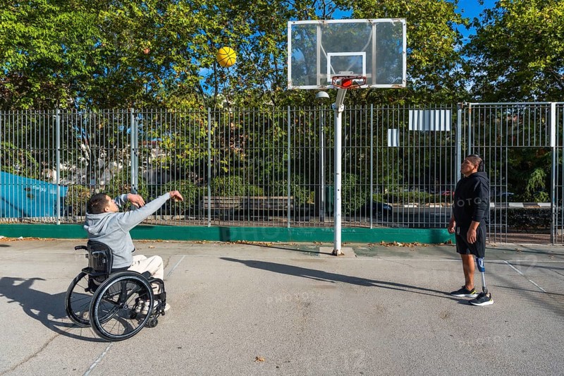 Two young men, one using a wheelchair and another with a prosthetic leg, playing basketball on an outdoor court, embracing inclusion and enjoying adaptive sports