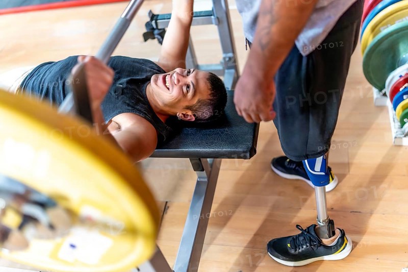 Smiling athlete on a bench press lifting weights while a man with a prosthetic leg stands by, offering support and encouragement during a challenging workout in a gym
