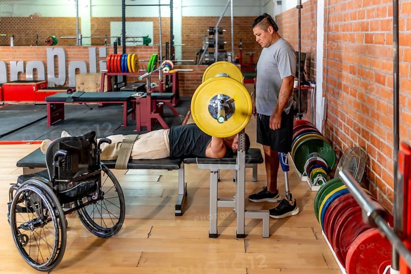Two determined men training in a gymone bench pressing weights while his teammate with a prosthetic leg stands supportivelyshowing adaptive strength and inclusion in sport