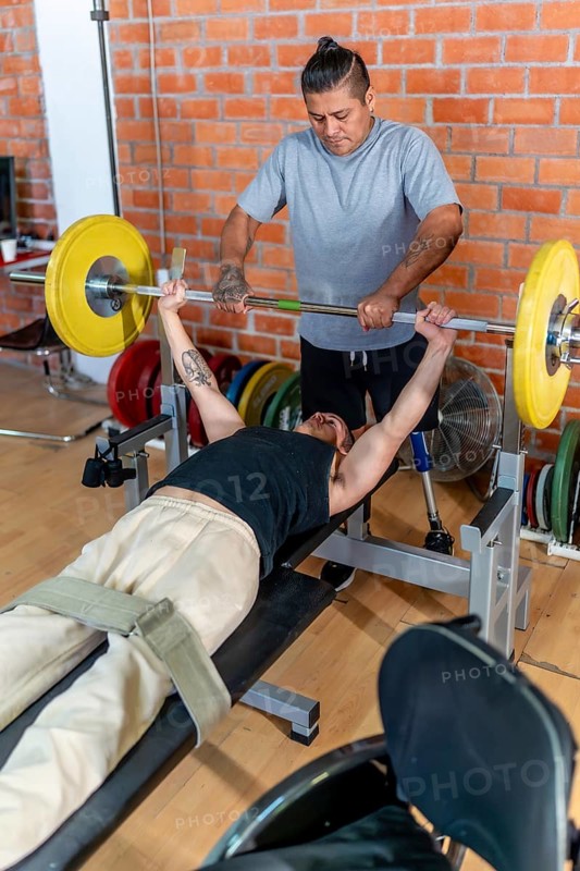 Young man with a prosthetic leg performing bench press exercises at the gym, receiving support and spotting from his personal trainer, focusing on strength and rehabilitation