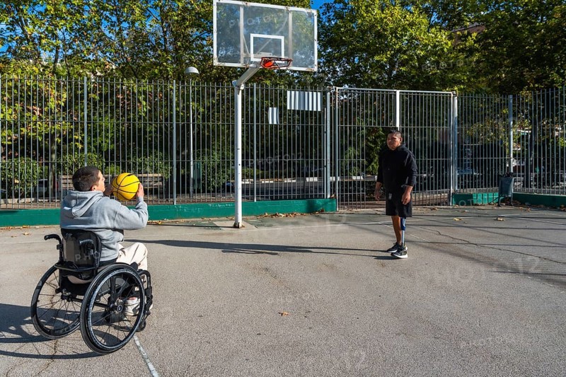 Two young men with disabilities, one using a wheelchair and the other a prosthetic leg, enjoying a game of basketball together on an urban court, promoting inclusion and adaptive sports