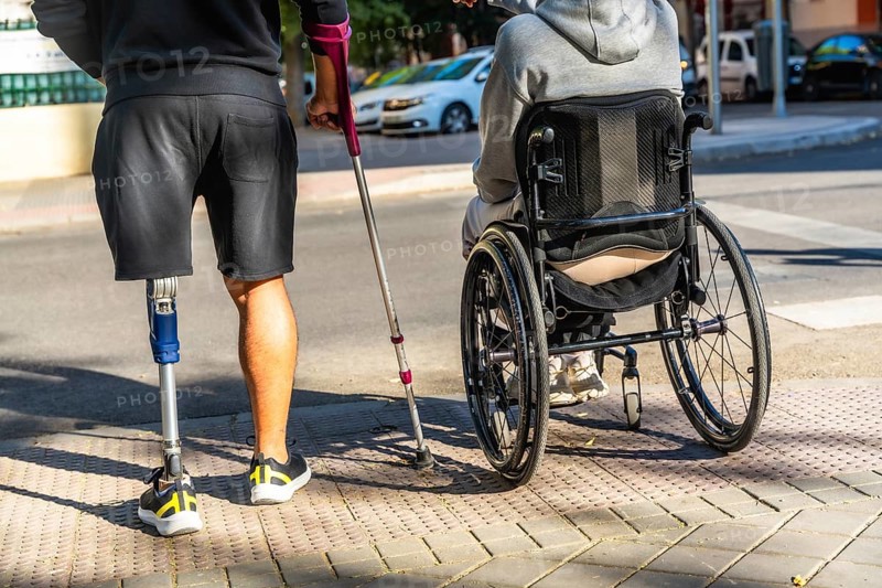 Two people with disabilities moving on a city street, one using a prosthetic leg and a crutch, and the other sitting in a wheelchair, representing friendship and inclusivity