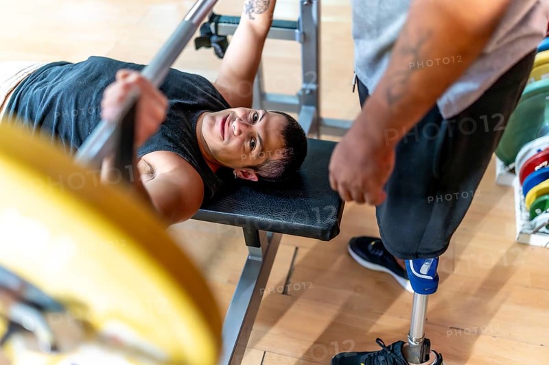 Disabled man performing a bench press with a barbell while a friend with a prosthetic leg stands by, offering support during a workout session at the gym