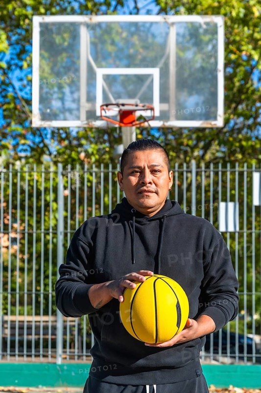 Indigenous man standing on an outdoor basketball court holding a bright yellow basketball, looking seriously at the viewer on a sunny day with a hoop in the background