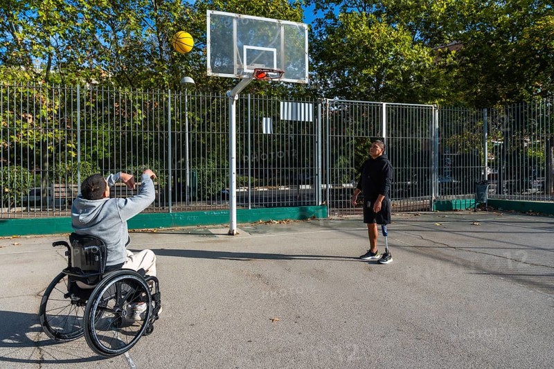 Two disabled friends are enjoying a game of basketball on an outdoor court, with one man in a wheelchair shooting the ball while his companion with a prosthetic leg watches
