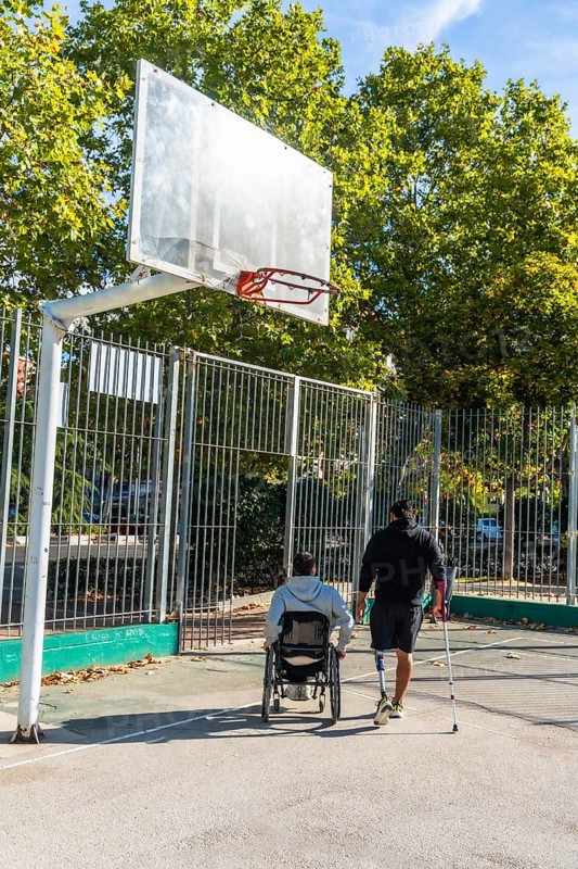Disabled men on an outdoor basketball court, one in a wheelchair and another with a prosthetic leg and crutch walking toward the hoop, showcasing inclusion and determination