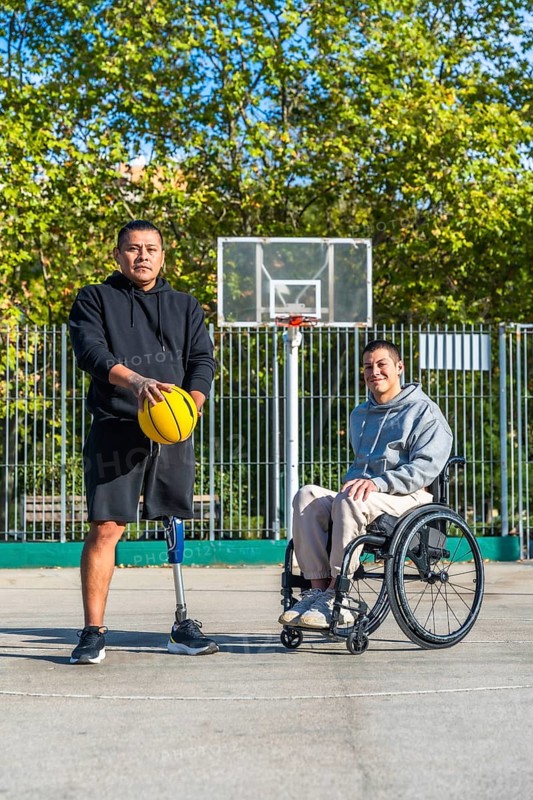 Two men, one with a prosthetic leg holding a basketball and another sitting in a wheelchair, standing together on an outdoor basketball court demonstrating inclusivity in sports