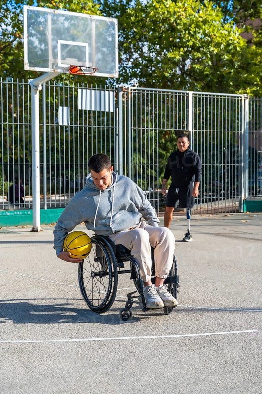 Young men with physical disabilities engaging in adaptive basketball. One person using a wheelchair and another with a prosthetic leg. Demonstrating inclusion and overcoming challenges through sports
