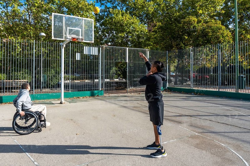 Two athletes playing basketball on an outdoor court. One sitting in a wheelchair and the other standing with a prosthetic leg. Demonstrating inclusion. Determination. And adaptive sports participation