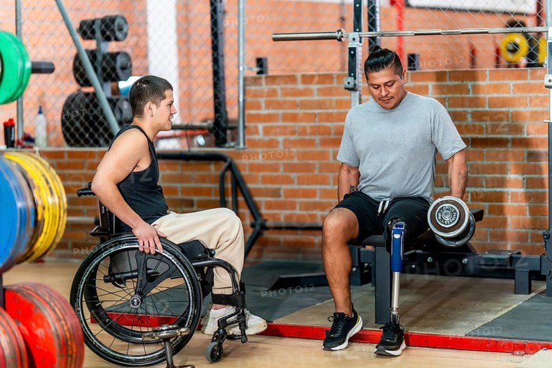 Two men with disabilities, one using a wheelchair and another with a prosthetic leg, engaging in fitness activities and supporting each other during a workout session at the gym