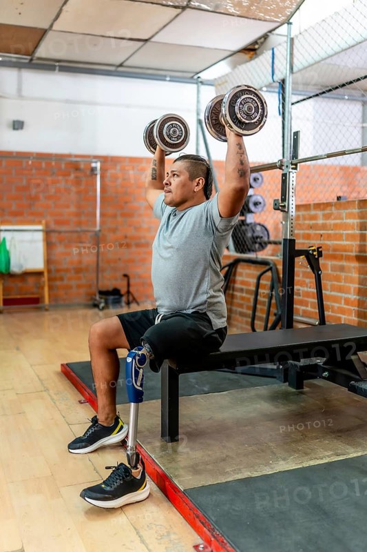 Man with a prosthetic leg performing overhead dumbbell presses while sitting on a weight bench, focusing on upper body strength and adaptive fitness in a gym environment