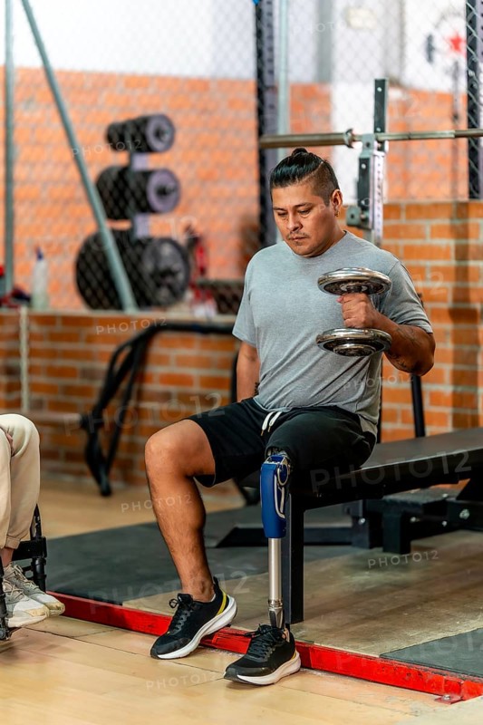 Disabled man with a prosthetic leg and an alternative hairstyle lifting a dumbbell in a gym, focusing on strength training and rehabilitation while sitting on a workout bench