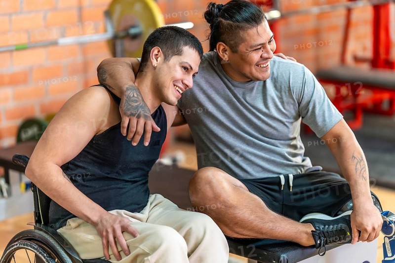 Two diverse adult men with disabilities smiling and embracing, one in a wheelchair and the other with a prosthetic leg, sharing a moment of joy and friendship at a gym