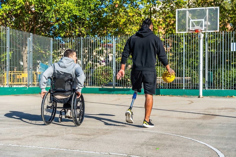 Man with prosthetic leg dribbling basketball on outdoor court while friend in a wheelchair watches, highlighting inclusion, adaptive sports, teamwork and active urban recreation
