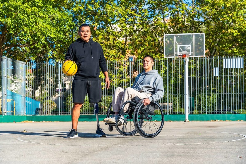 Two young men, one with a prosthetic leg holding a basketball and another sitting in a wheelchair, are standing together on an outdoor basketball court, representing inclusion and adaptive sports
