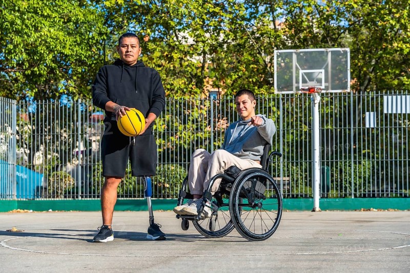 Men with diverse disabilities, one using a prosthetic leg and holding a basketball, the other sitting in a wheelchair and pointing towards the viewer, posing on an outdoor basketball court