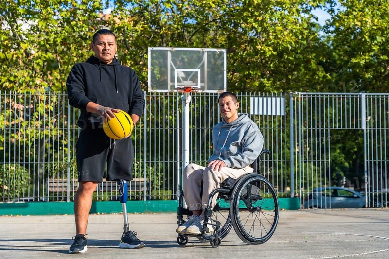 Two young men, one with a prosthetic leg holding a basketball and another sitting in a wheelchair, standing together on an outdoor basketball court in a park, representing adaptive sports