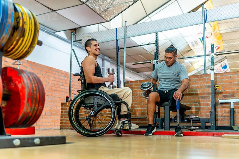Two men with disabilities training together in a gymone in a wheelchair, one with a prosthetic leglifting dumbbells, showing strength, determination, teamwork and inclusion