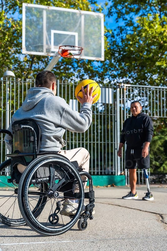 Man in a wheelchair aiming to shoot a basketball while his male friend, who uses a prosthetic leg, stands watching on an outdoor basketball court, representing inclusion and adapted sports