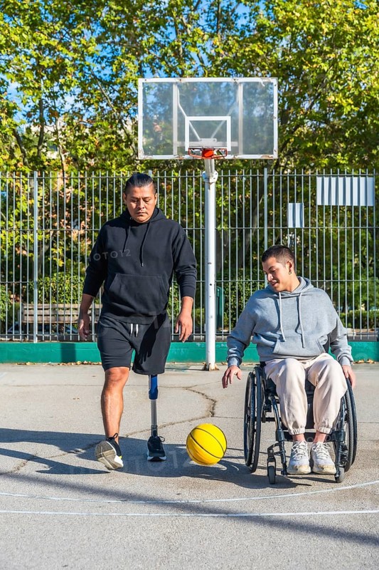 Two friends play basketball on an outdoor court, one man with a prosthetic leg, the other in a wheelchair dribbling a yellow ball, celebrating inclusion and active friendship